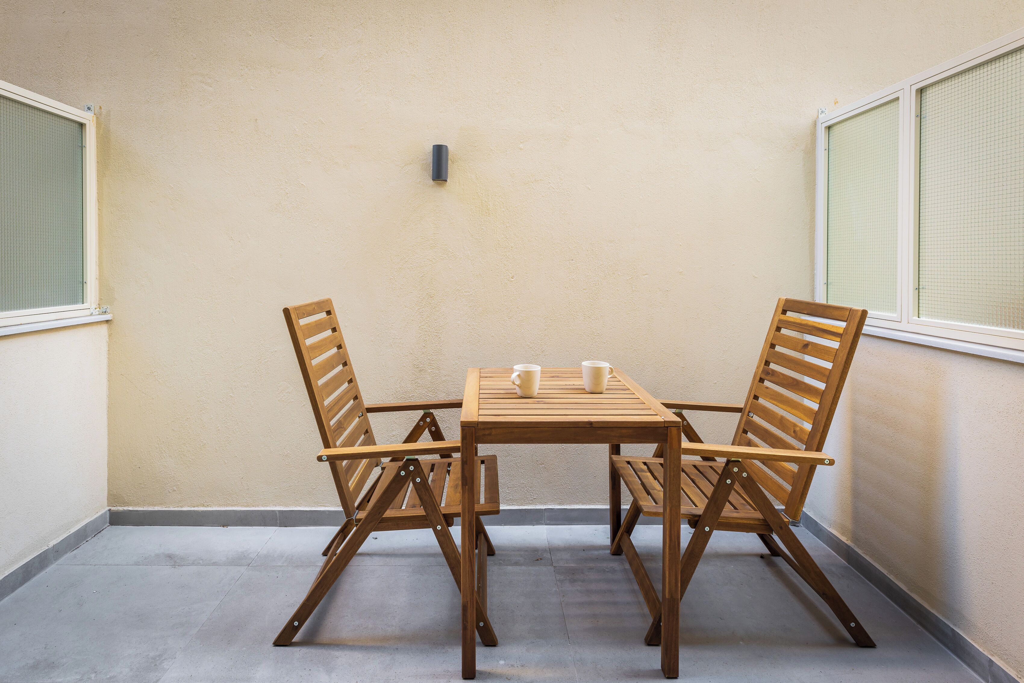 Un balcón pequeño y cerrado con una mesa de madera y dos sillas, una lámpara pequeña fijada a la pared y una ventana con mosquitera.