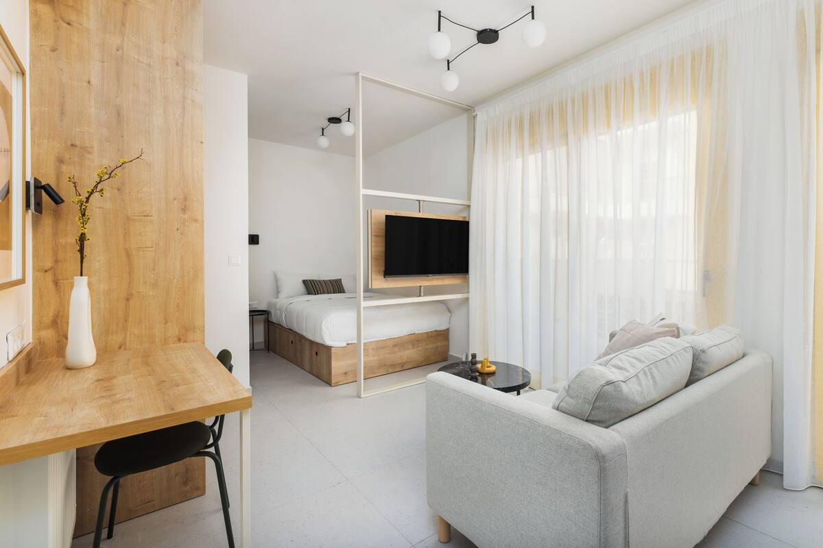 A modern, well-lit room with a wooden desk, a grey sofa, a bed with a TV, and a large window with sheer curtains.