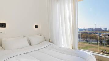 A bedroom with a large bed, white bedding, a window with a view of a cityscape, and a curtain.