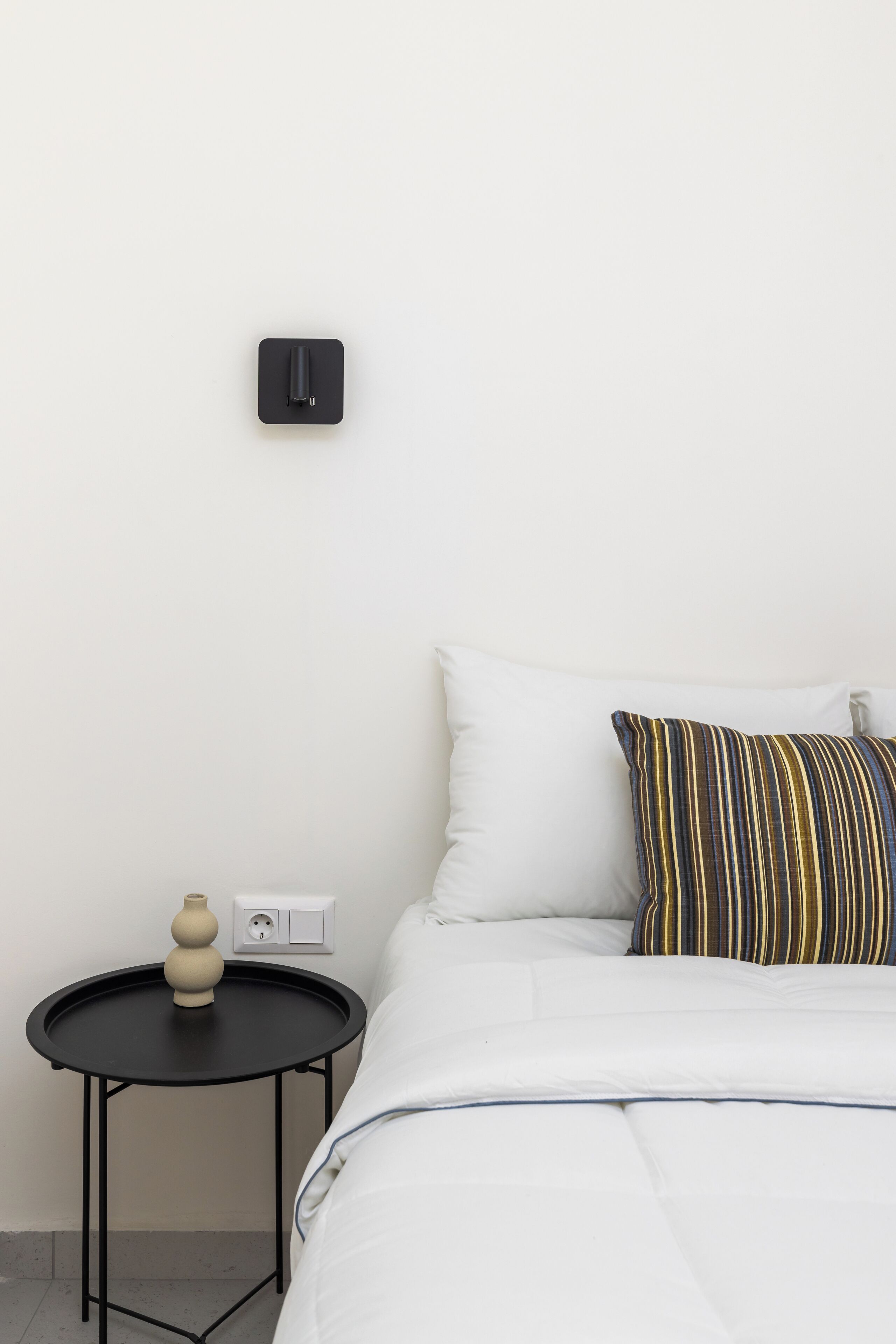 Un lit bien fait, avec une couette blanche et un oreiller à rayures, une petite table noire sur laquelle se trouve un vase, et une prise électrique au mur.