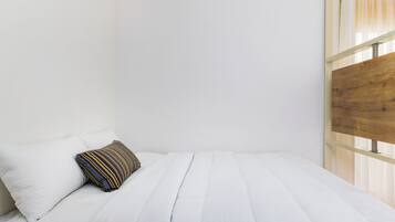 Una cama individual con ropa de cama blanca y un almohadón a rayas, cabecero de madera y una ventana con cortinas traslúcidas.