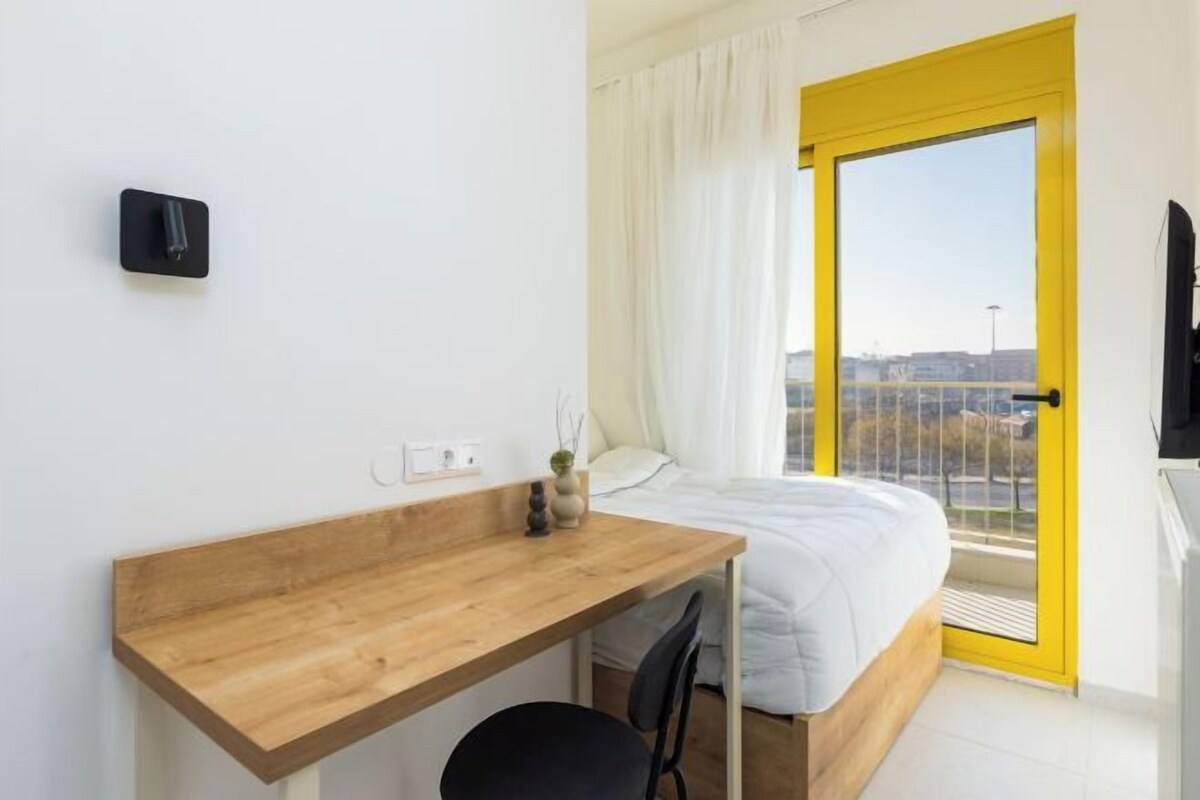 Un dormitorio moderno con un escritorio de madera, una cama, un ventanal con detalles amarillos y vistas a la ciudad.