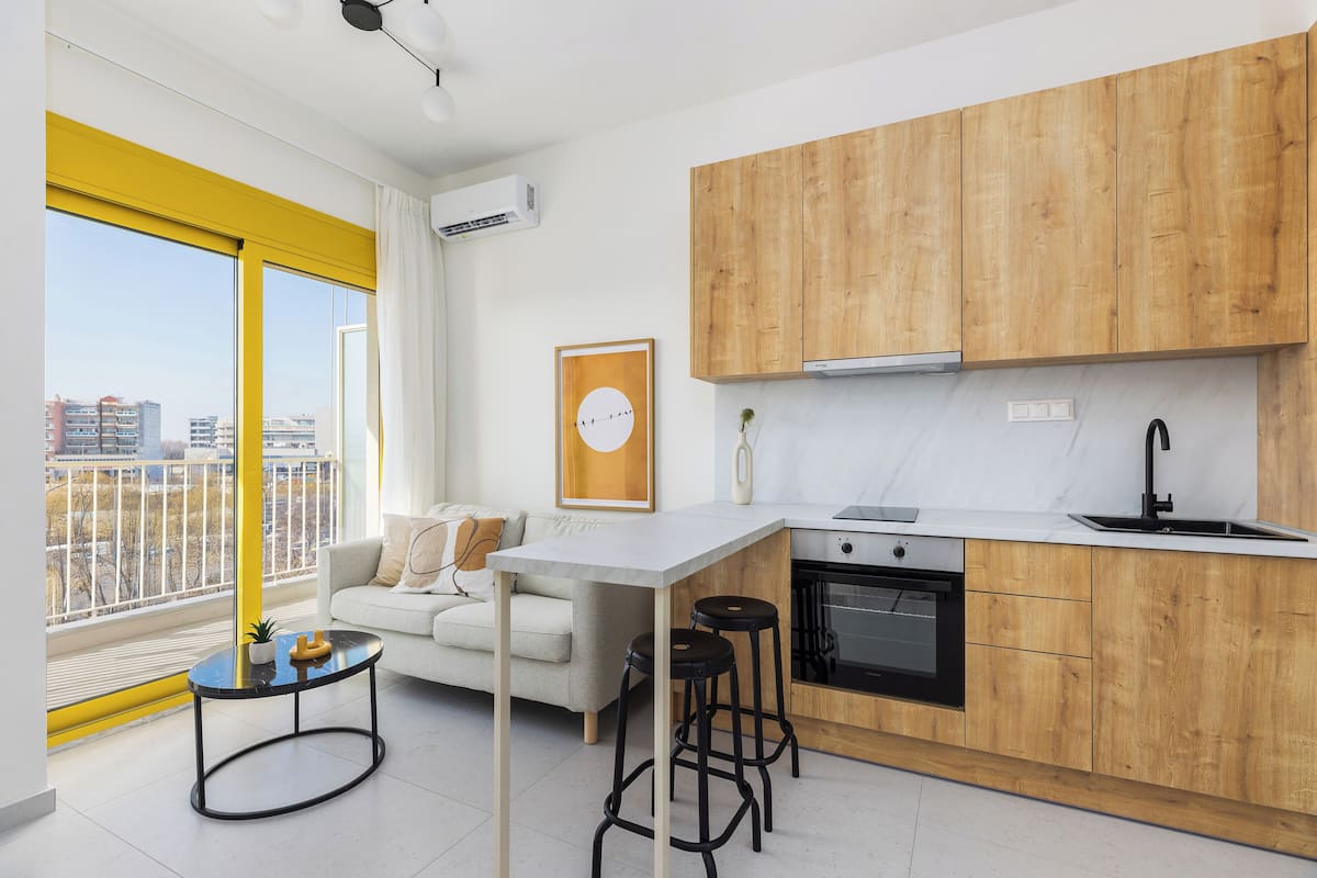 A modern kitchen with wooden cabinets, a white countertop, and a built-in oven. There is a small round table with stools in the living area.