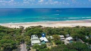 An aerial view of a coastal area with a beach, dense greenery, and several buildings with distinct roofs. - Driftwood on Home Beach - Absolute Beachfront (Point Lookout)