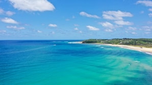 Un paysage côtier avec une mer bleue et limpide, une plage de sable et des collines verdoyantes en arrière-plan.
