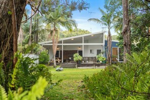 Une maison à deux étages dotée d’un patio couvert, entourée d’une végétation luxuriante et d’arbres.