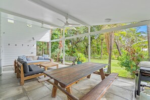 Une terrasse extérieure couverte, avec une table et des bancs en bois, un canapé avec des coussins et un barbecue.