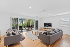 A modern living room with a sofa set, a coffee table, a flat-screen TV, and a sliding glass door leading to an outdoor patio. - Driftwood on Home Beach - Absolute Beachfront (Point Lookout)