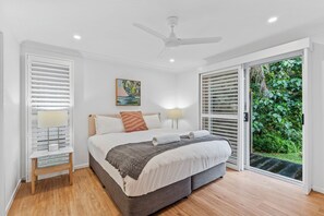 Une chambre à coucher avec un lit, des lampes de chevet, un ventilateur de plafond et une vue sur la verdure, visible à travers la porte-fenêtre coulissante.