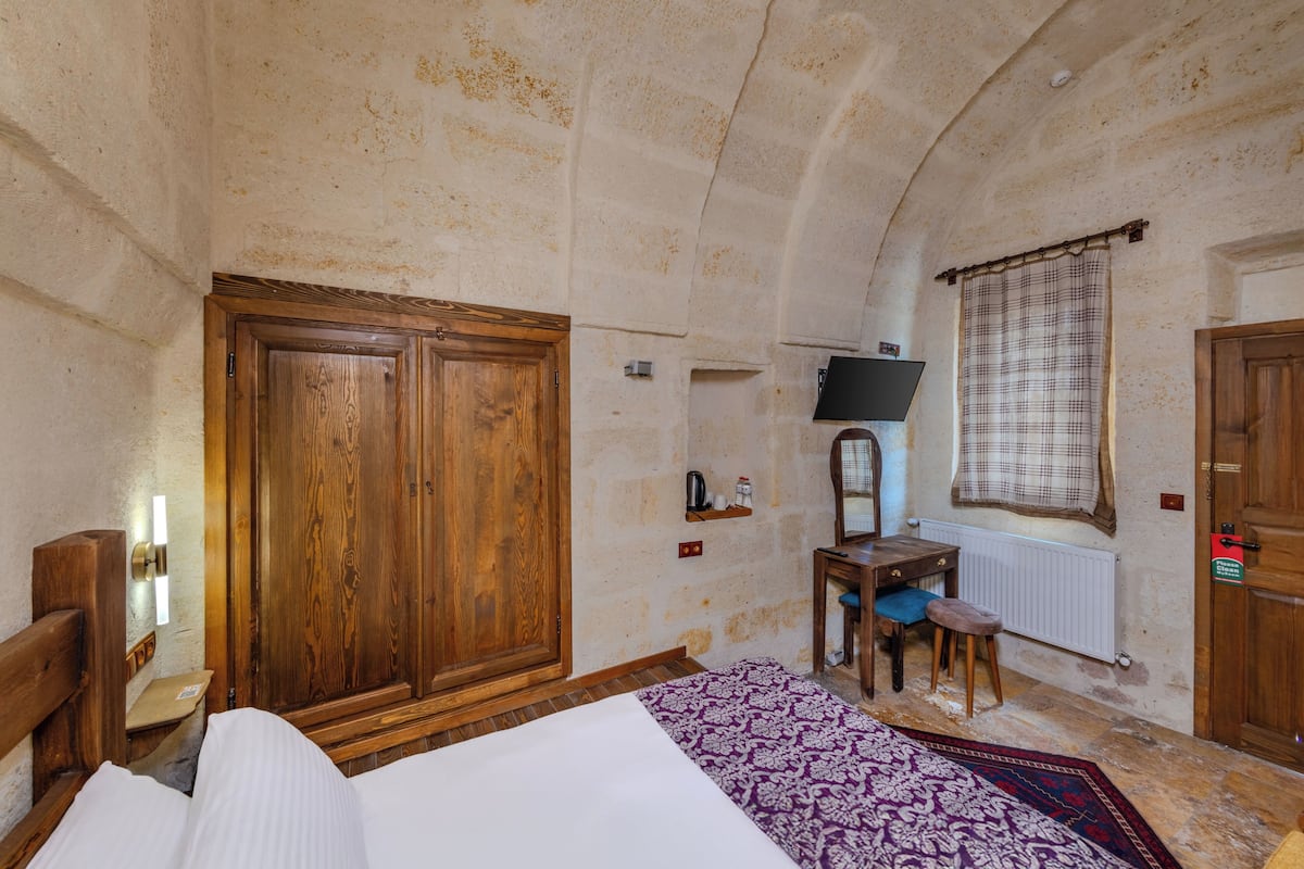 A room with a bed, wooden wardrobe, a small table with a chair, a mirror, and a window with a checkered curtain.