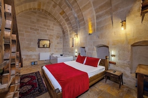 A stone-walled bedroom with a wooden bed, a red blanket, a wooden nightstand, a small table, a wooden ladder, a bathroom with a bathtub, and a red rug.