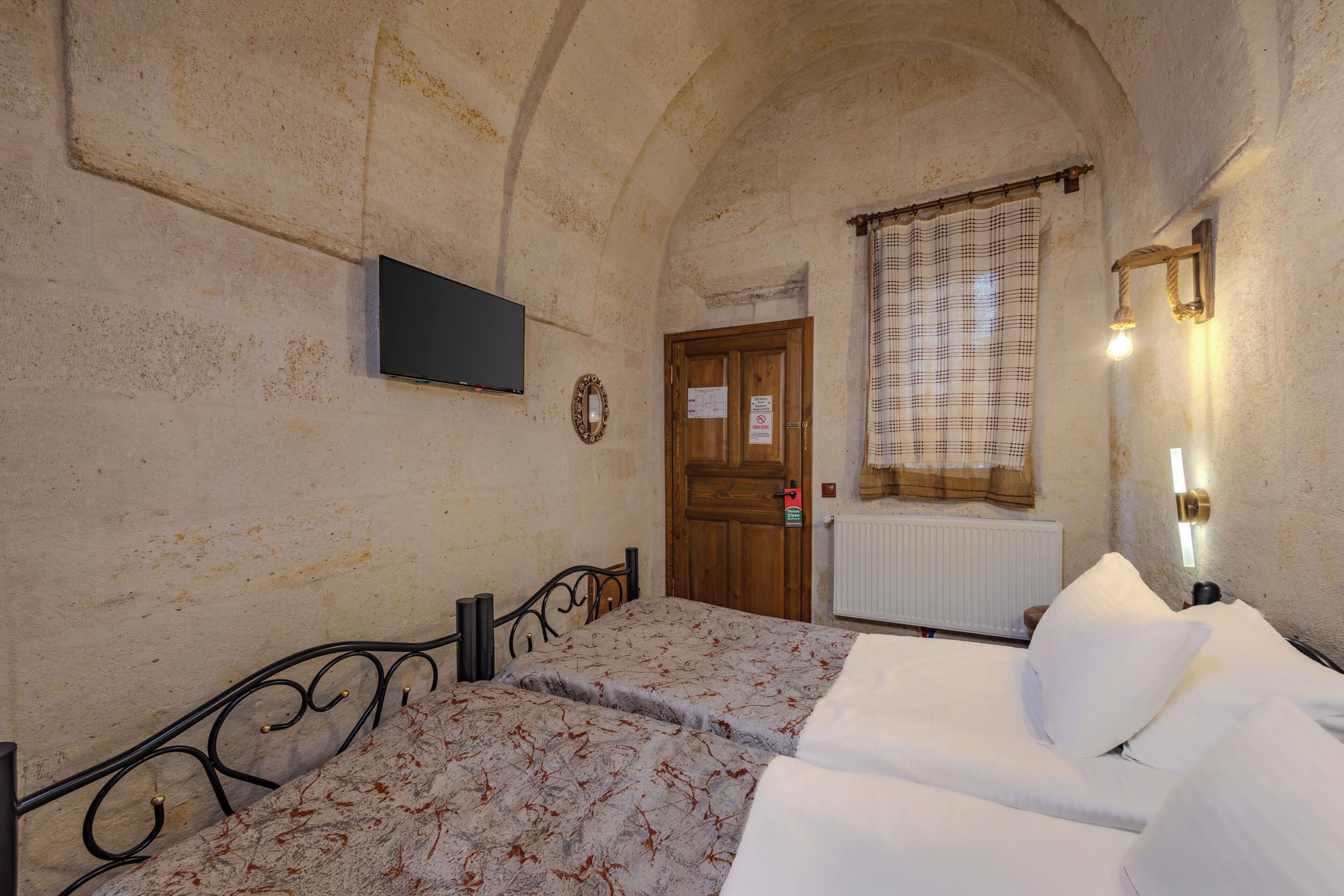 A room with a bed, a television mounted on the wall, a wooden door, and a window with a curtain.