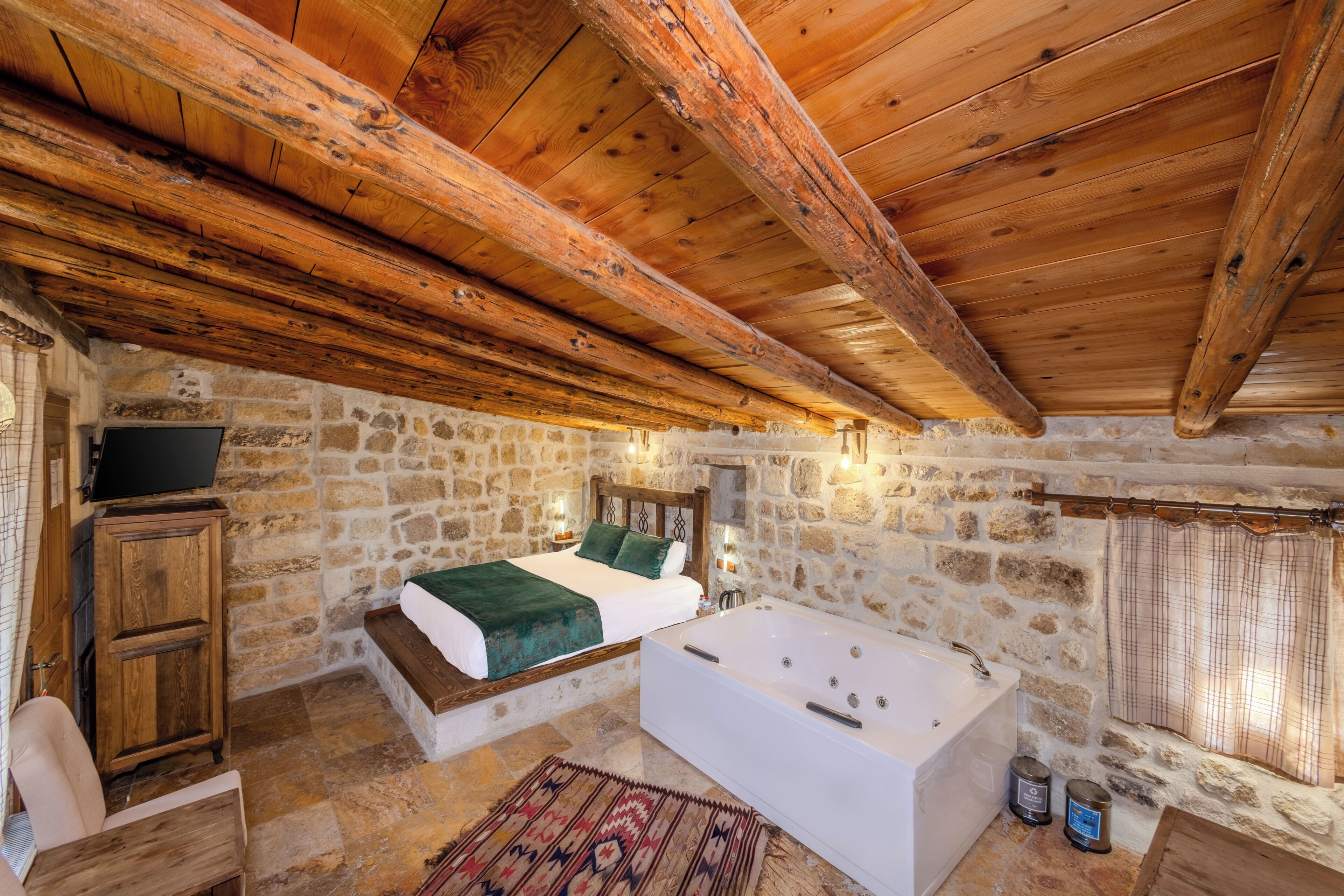 A bedroom with a stone wall, wooden ceiling beams, a bed, a bathtub, and a wooden cabinet.
