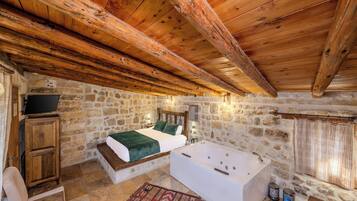 A bedroom with a stone wall, wooden ceiling beams, a bed, a bathtub, and a wooden cabinet.