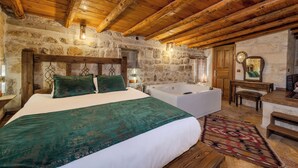 Una habitación rústica con pared de piedra, vigas de madera en el techo, una cama grande con ropa de cama verde, una bañera independiente y un banco de madera.