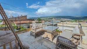Una terraza en la azotea con mobiliario de mimbre, una mesa de vidrio y vistas a un paisaje urbano.