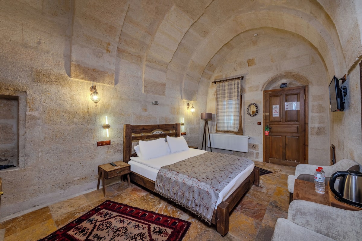 A room with a bed, a wooden nightstand, a table with a kettle, a water bottle, and a door with a sign.