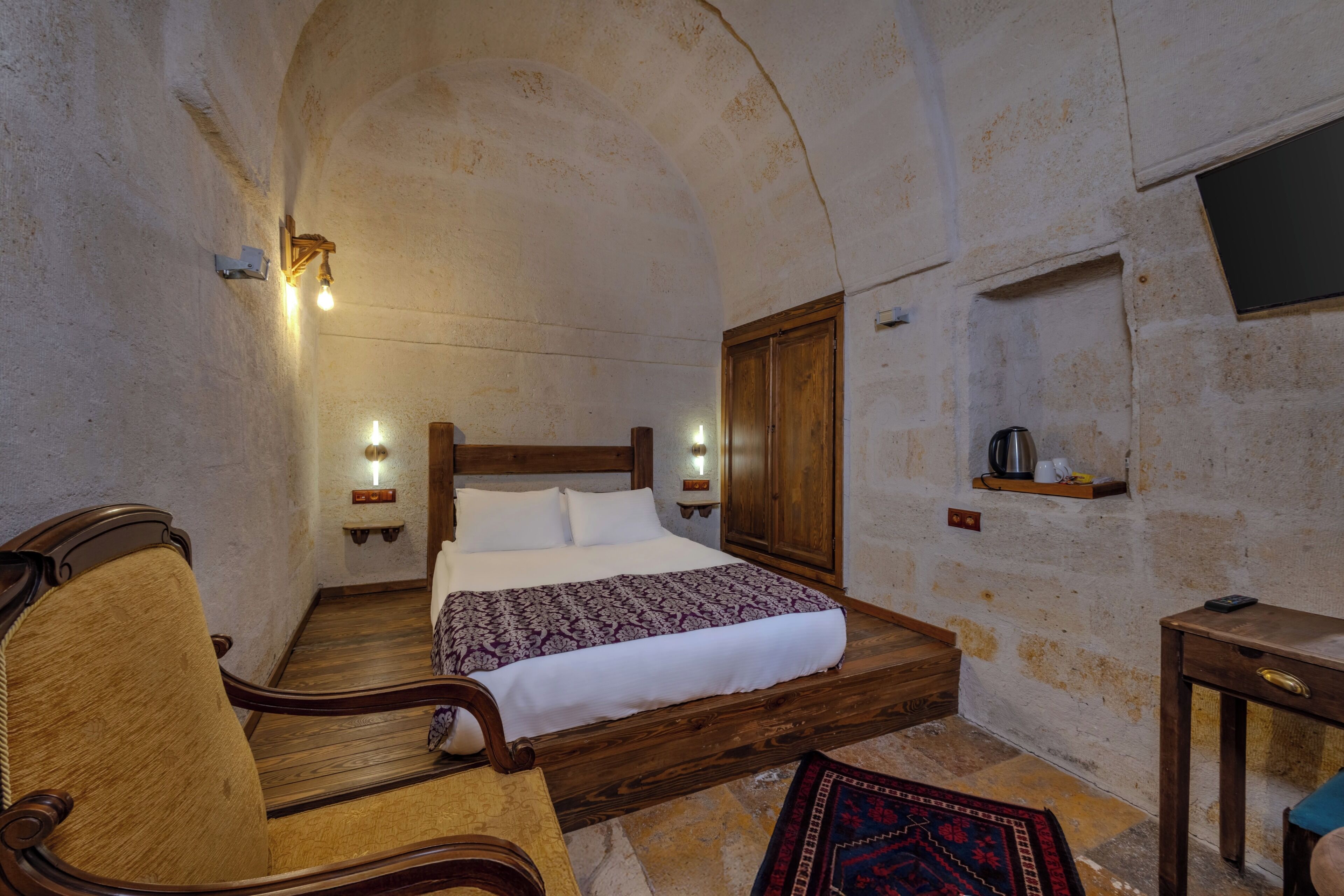A bedroom with a wooden bed, a chair, a small table, and a shelf with a kettle.