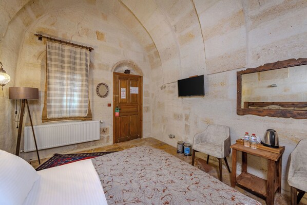 A room with a bed, a wooden table with bottles, a chair, a television, a radiator, and a window with a curtain. - Cappadocia Balloon House (Urgup)