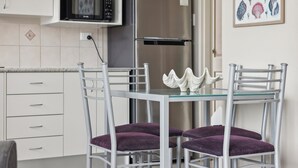 A modern kitchen with white cabinets, a stainless steel refrigerator, a microwave, and a glass dining table with two purple cushioned chairs. - Ocean Parade 18 - Two Bedroom Apartment with Pool Views (COFFS HARBOUR)