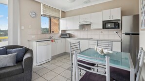 A compact kitchen with white cabinets, a microwave, and a dishwasher. There's a dining table with chairs and a sofa in the living area. - Ocean Parade 18 - Two Bedroom Apartment with Pool Views (COFFS HARBOUR)