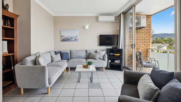 A modern living room with a sectional sofa, a coffee table, and a TV. There is a bookshelf and a balcony with a view. - Ocean Parade 18 - Two Bedroom Apartment with Pool Views (COFFS HARBOUR)