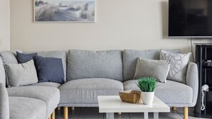 A modern living room with a grey sectional sofa, a white coffee table, a framed landscape painting, and a flat-screen TV. - Ocean Parade 18 - Two Bedroom Apartment with Pool Views (COFFS HARBOUR)
