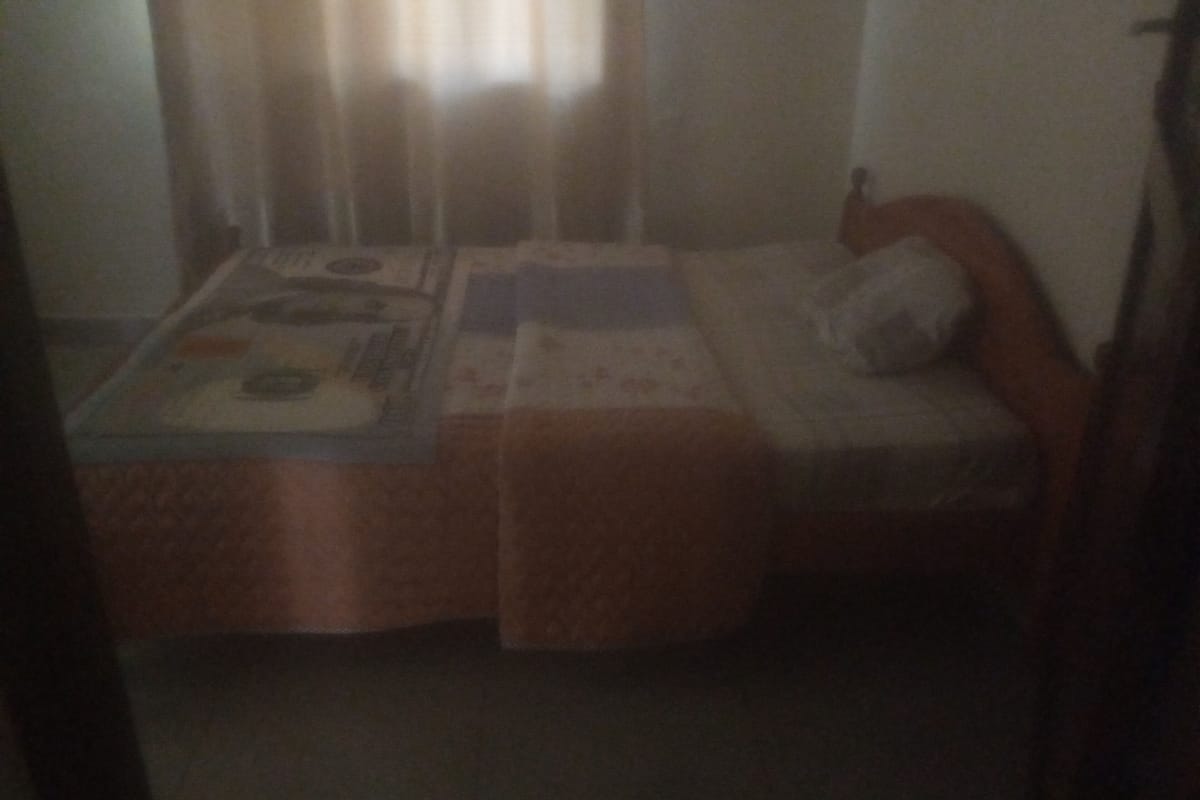 A small, dimly lit room with a single bed, a nightstand, and a window with curtains.