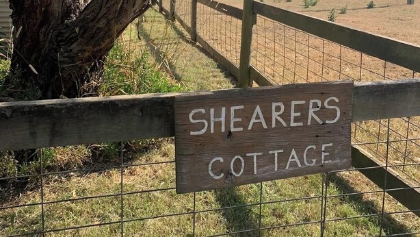 Ein hölzernes Schild mit der Aufschrift 'Shearers Cottage', mit einem eingezäunten Bereich und Bäumen im Hintergrund.