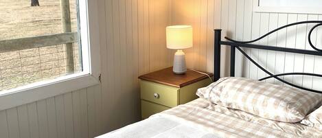 A bedroom with a bed, a nightstand with a lamp, a window with a view of trees, and a framed picture on the wall.