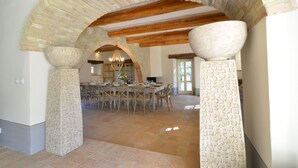 Un comedor espacioso con un techo alto y vigas de madera a la vista, columnas de piedra con intrincados relieves, y una lámpara de araña grande.