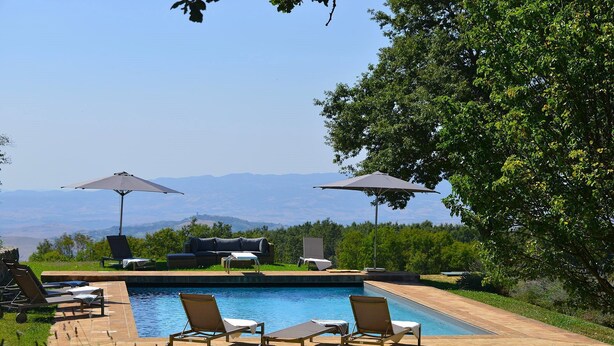 Zona de piscina con sillas de descanso y sombrillas, rodeada de vegetación y vistas a montañas distantes.