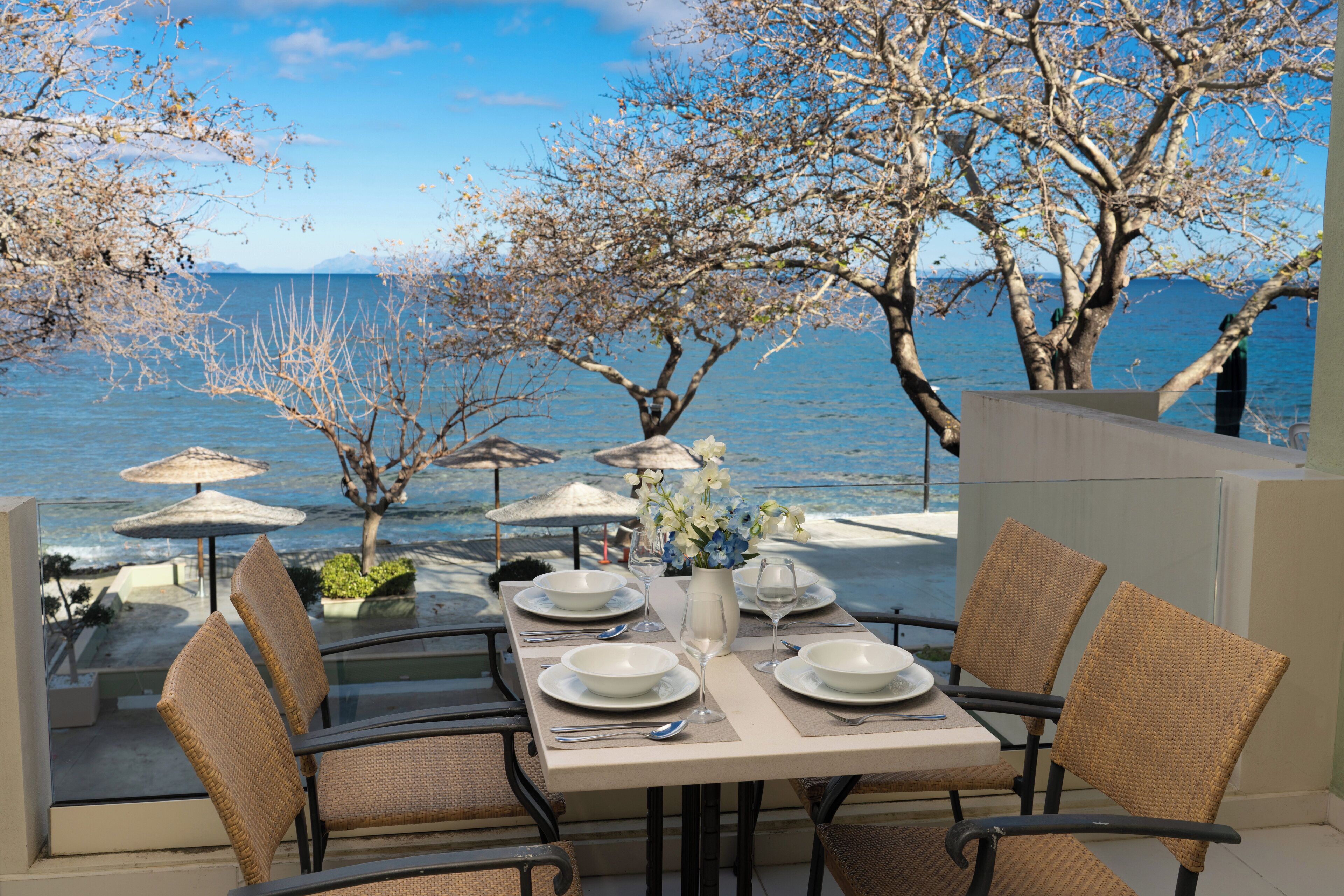 A dining table set with white plates, silverware, and a vase of flowers, overlooking a body of water with trees and umbrellas.