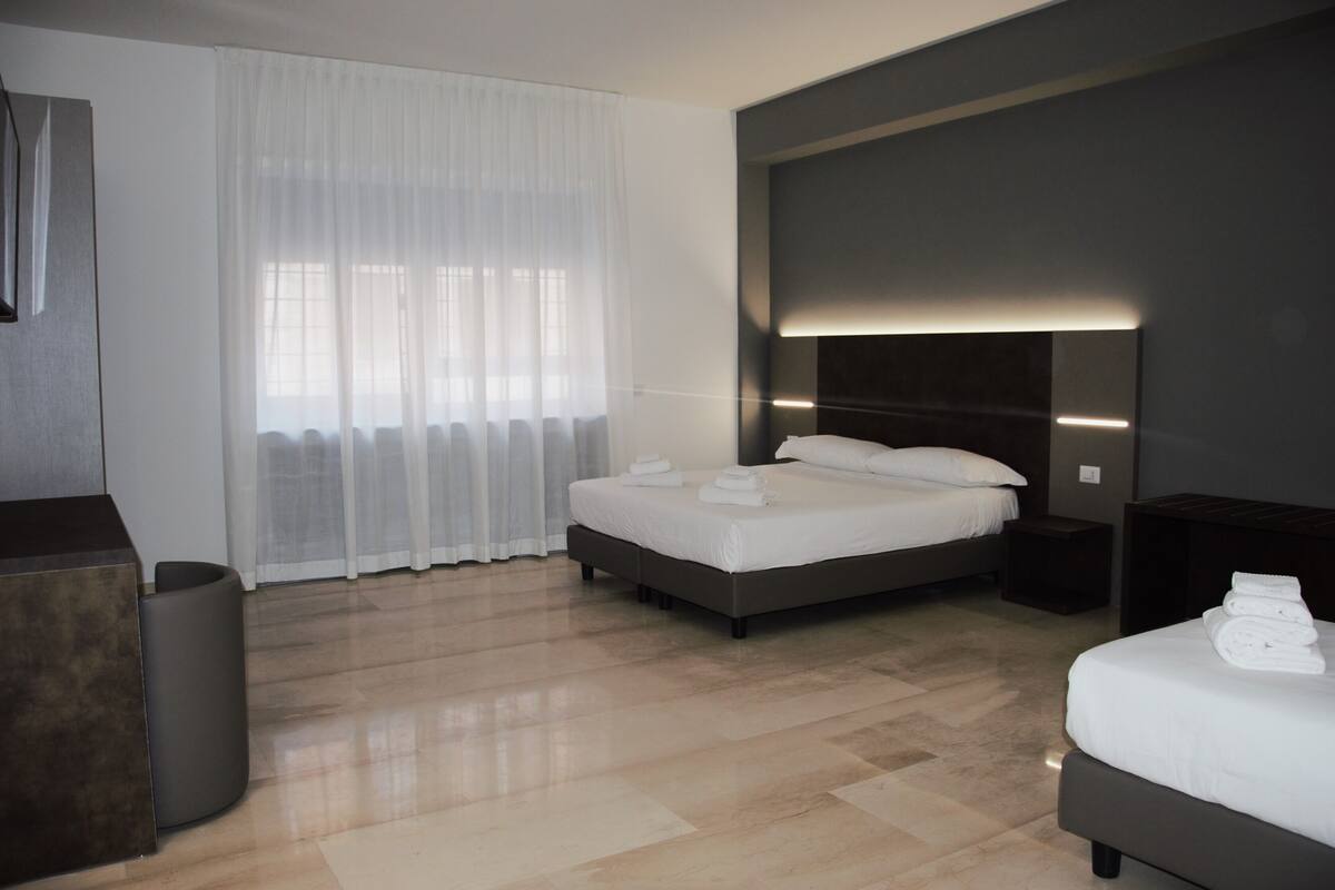 A modern hotel room with a large bed, bedside tables, a bedside lamp, and a view of the city through sheer curtains.