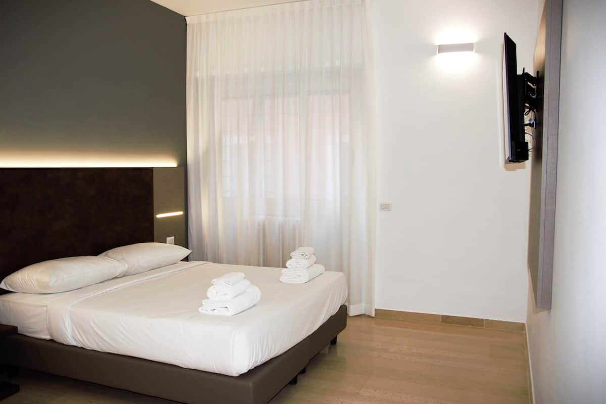 A neatly made bed with white linens and towels, a dark headboard, a large window with sheer curtains, and a wall-mounted TV.