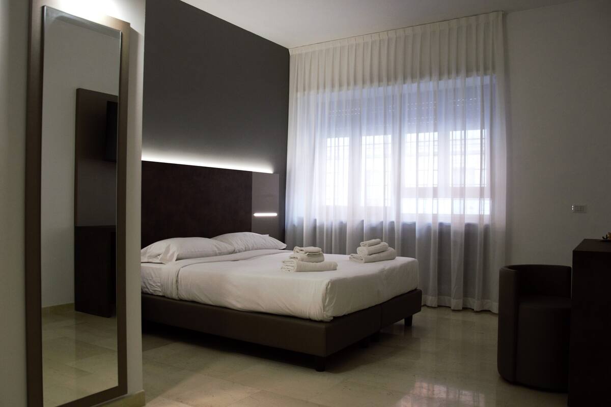 A modern hotel room with a large bed, a dark headboard, a large mirror, and a view of the outdoors through sheer curtains.