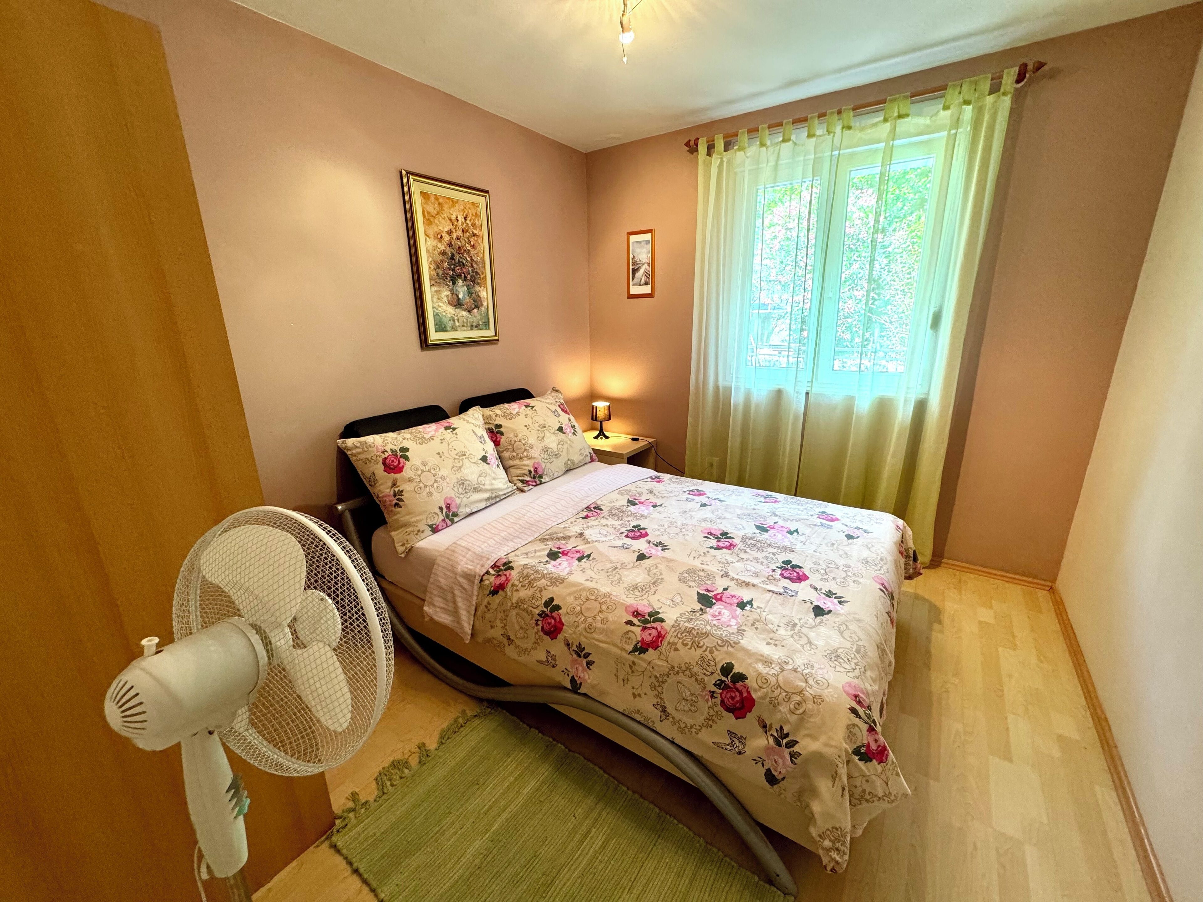 A bedroom with a bed, a standing fan, a green rug, and a window with curtains.