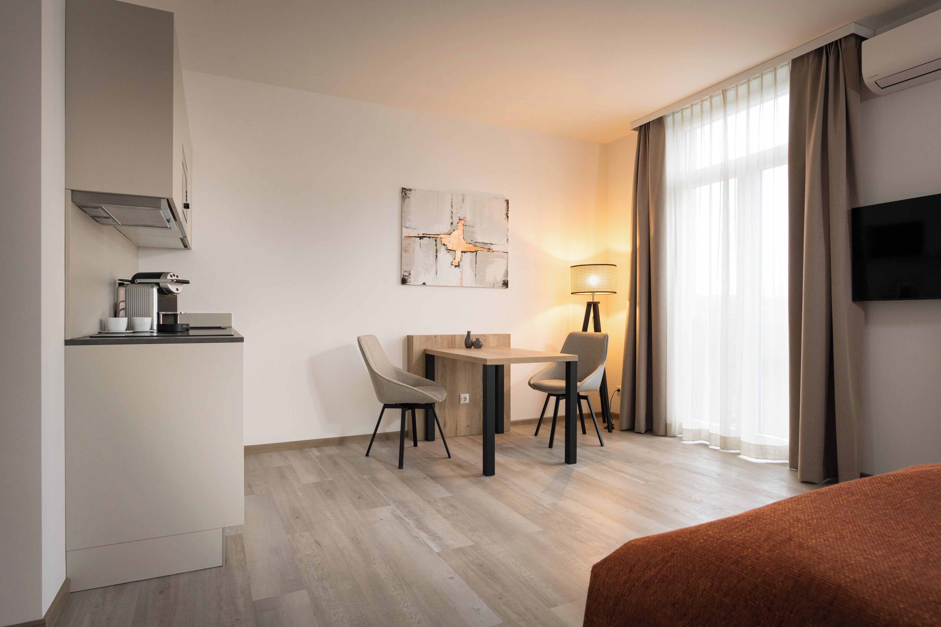 A hotel room with a bed, a desk with chairs, a TV, a lamp, and a painting on the wall.