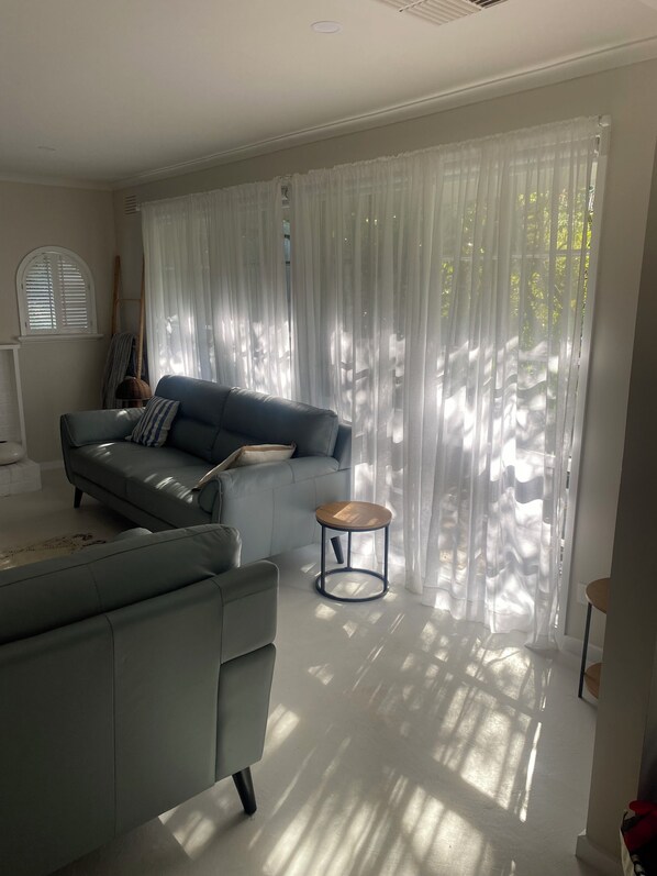 A modern living room with a grey sofa, a round wooden side table, and sheer white curtains. - Cosy Beach cottage 500 meters to Blairgowrie front beach peaceful and private. (Blairgowrie)