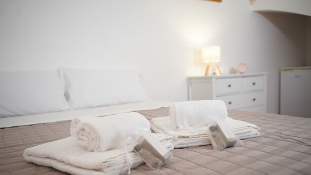 Un lit bien fait, avec des serviettes blanches pliées en rouleau et une table de chevet blanche sur laquelle se trouve une lampe.