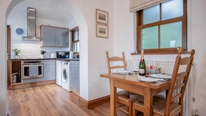 A compact kitchen with modern appliances, a dining area with wooden furniture, and a window with frosted glass. - Marina House Annex - 1 Bedroom - Neyland (Milford Haven)
