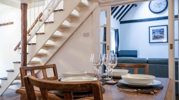 A dining area with a wooden table set for four, including plates, glasses, and cutlery. A wooden chair is positioned in front of the table. A staircase with wooden railings is visible to the left. - Stable Cottage - 1 Bedroom Cottage - Penally (Tenby)