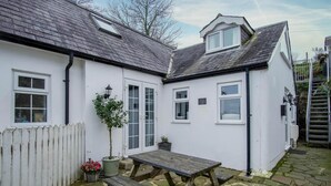 A white cottage with a stone patio, a wooden table, and a small garden. - Stable Cottage - 1 Bedroom Cottage - Penally (Tenby)