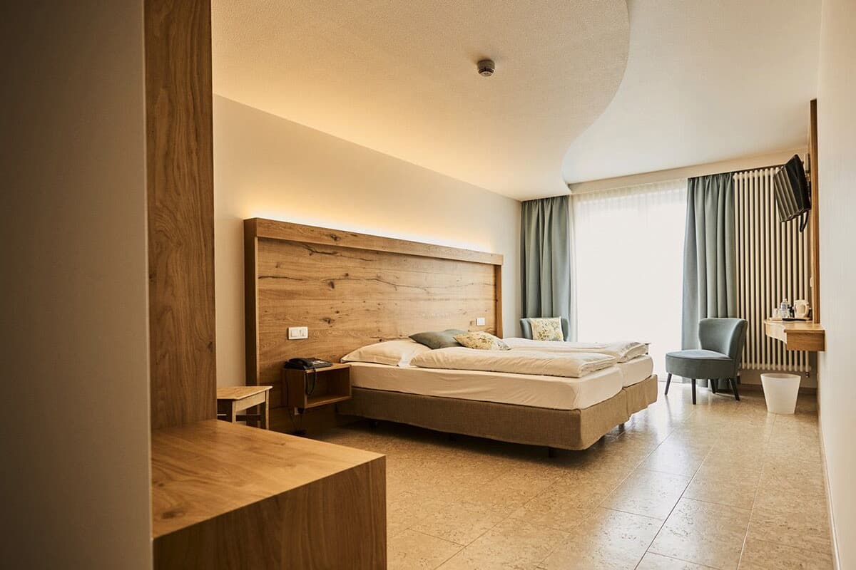 Una habitación de hotel moderna con una cama grande, mesita de noche, silla y un pequeño escritorio con lámpara.