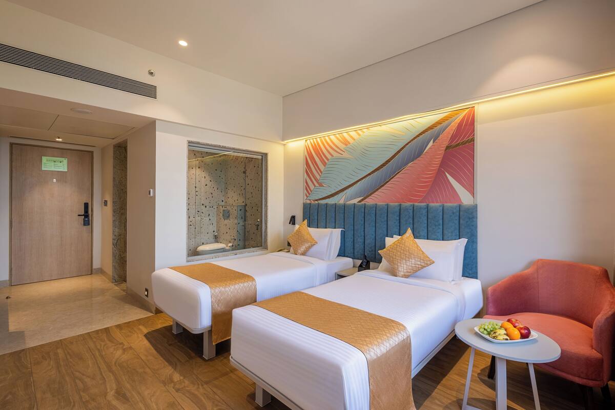A hotel room with two beds, a small table with fruit, a red chair, and a large abstract painting on the wall.