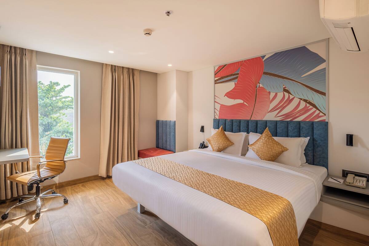 A hotel room with a large bed, a desk, and a chair. There is a window with curtains, a painting on the wall, and a nightstand with a phone.