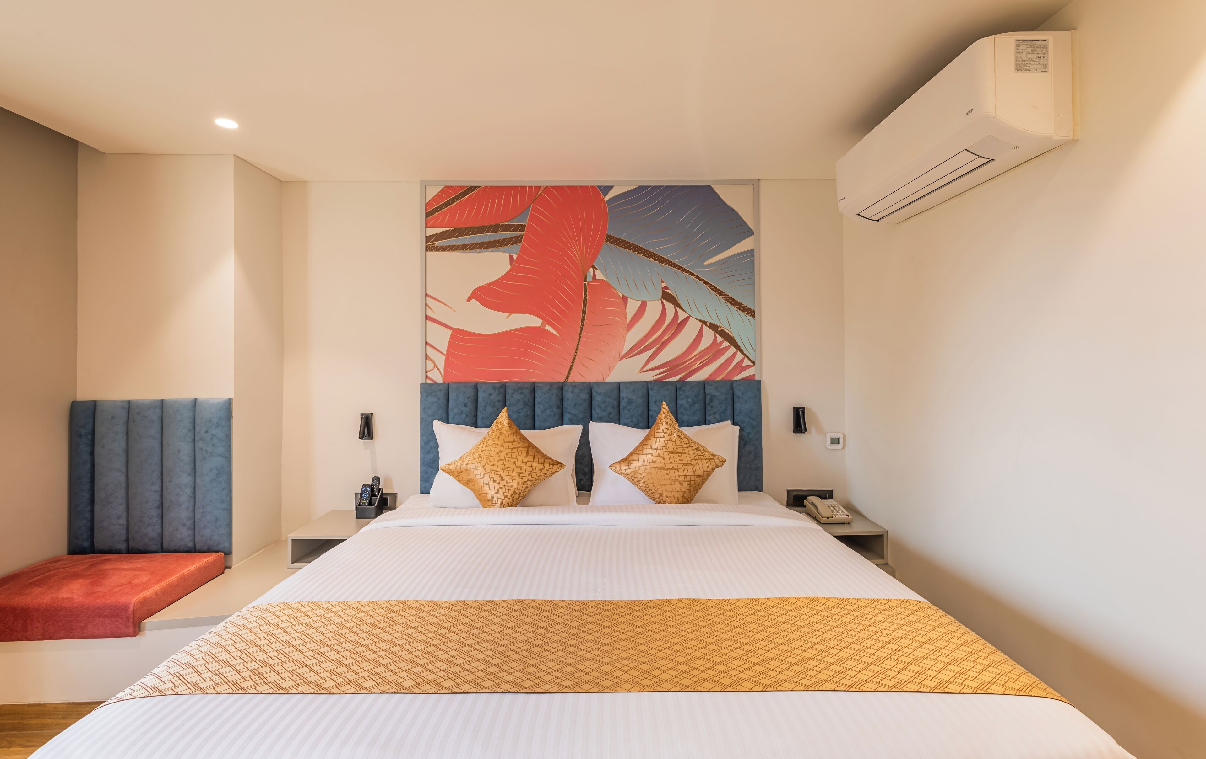 A modern hotel room with a large bed, a headboard with a blue panel, a red bench, and a wall-mounted air conditioner.