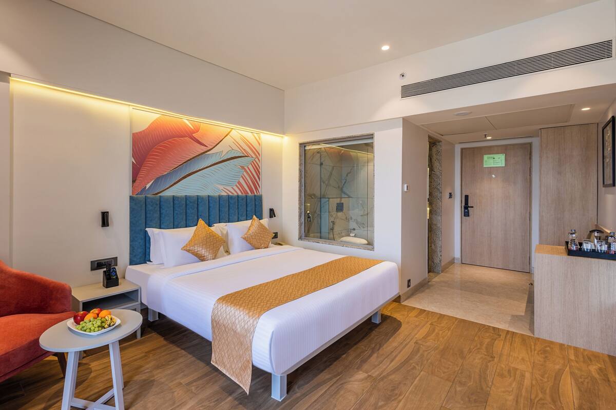 A modern hotel room with a large bed, a seating area with a red chair, a small table with fruit, and a bathroom with a glass shower enclosure.