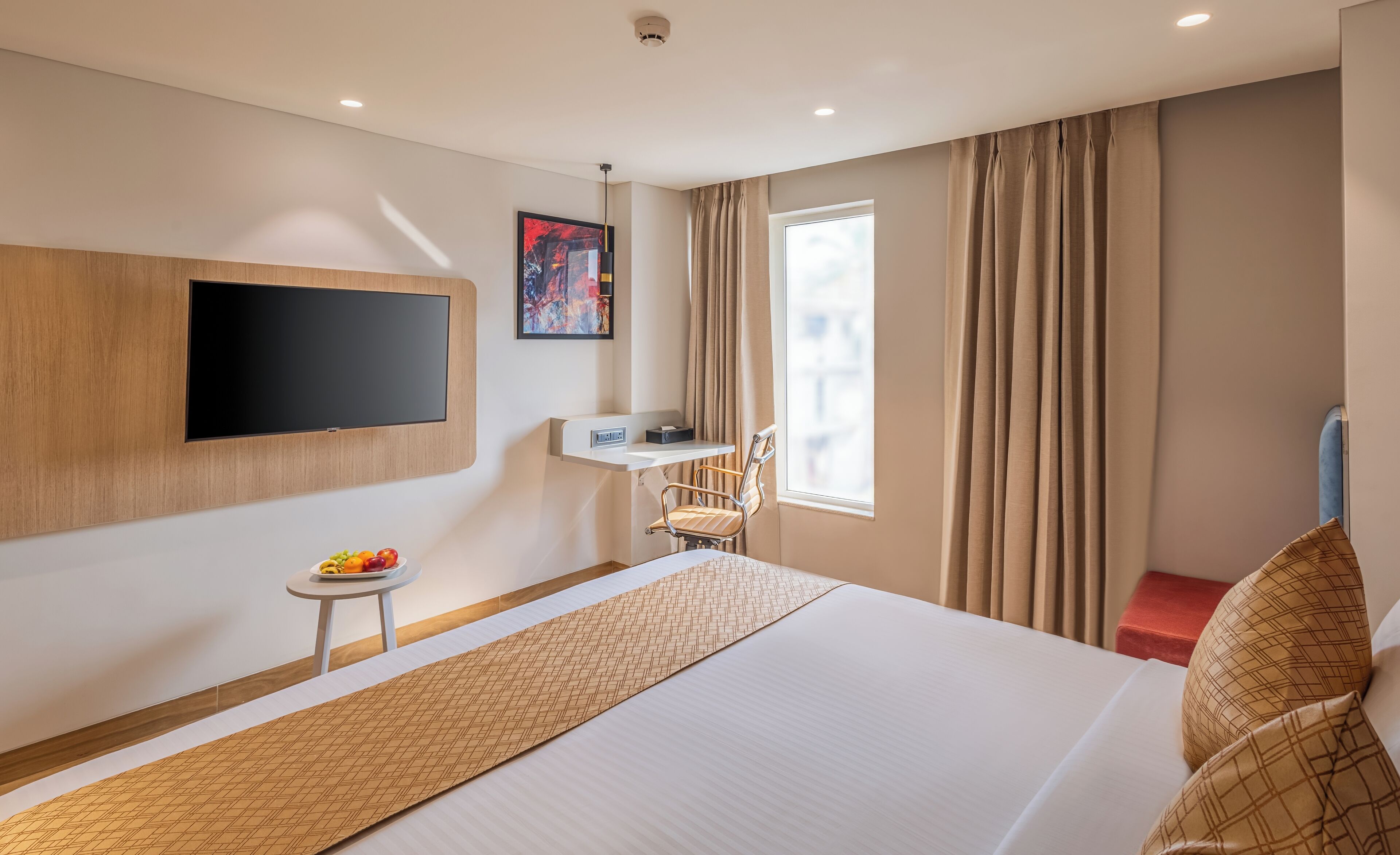 Habitación de hotel con una cama grande, un televisor de pantalla plana montado en la pared, una mesita con fruta, una silla y una ventana con cortinas.