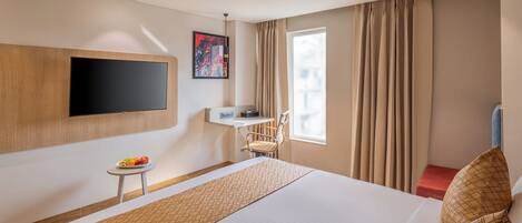 Habitación de hotel con una cama grande, un televisor de pantalla plana montado en la pared, una mesita con fruta, una silla y una ventana con cortinas.